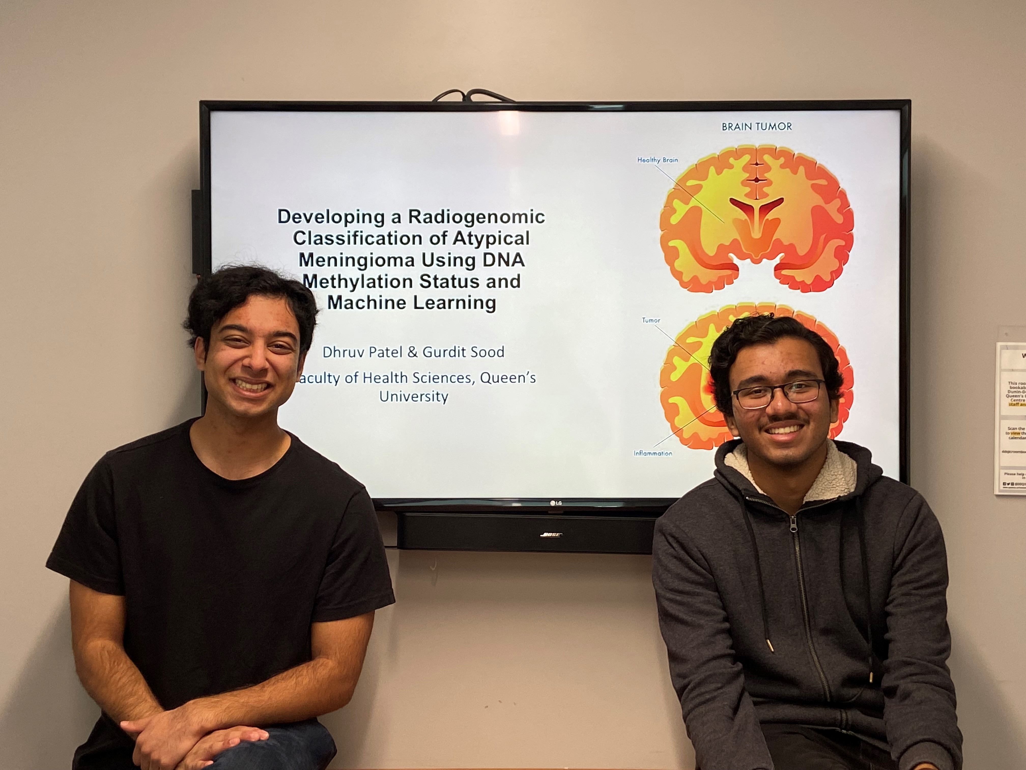 Gurdit Sood (left) and Dhruv Patel (right) sit in front of their scientific poster which is displayed on a TV screen.