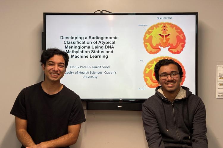 Gurdit Sood (left) and Dhruv Patel (right) sit in front of their scientific poster which is displayed on a TV screen.