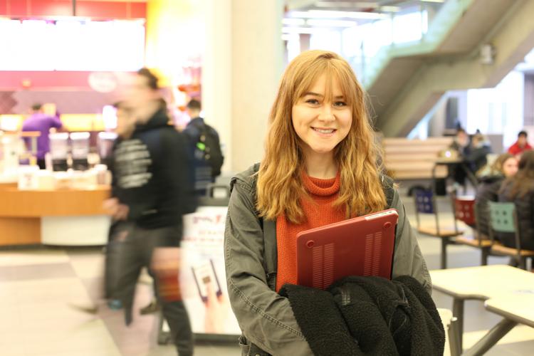 Young woman with red hair in a green jacket standing in the cafeteria while holding a red laptop.