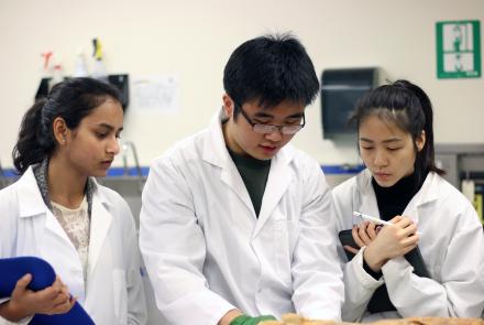 three students wearing white lab coats examine a specimen located just outside the bottom of the frame.